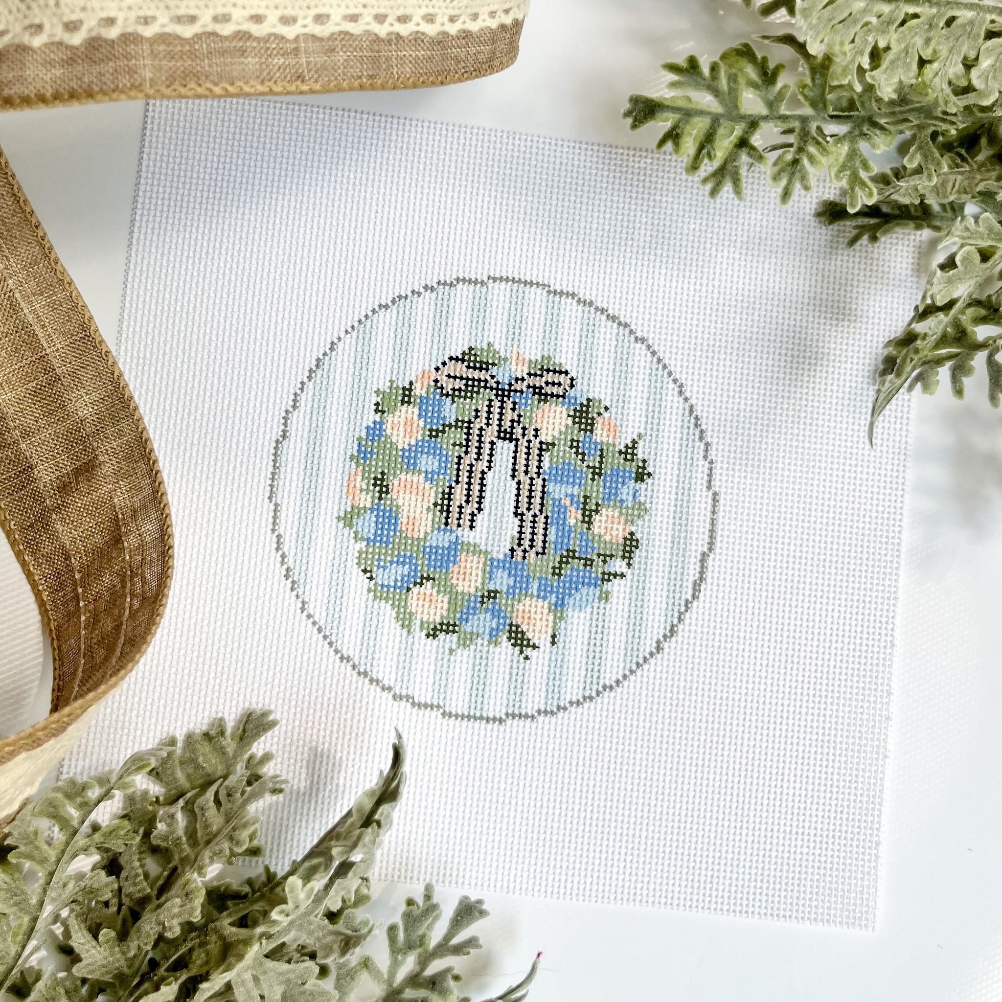 A needlepoint canvas featuring a green wreath with blue flowers and a striped bow